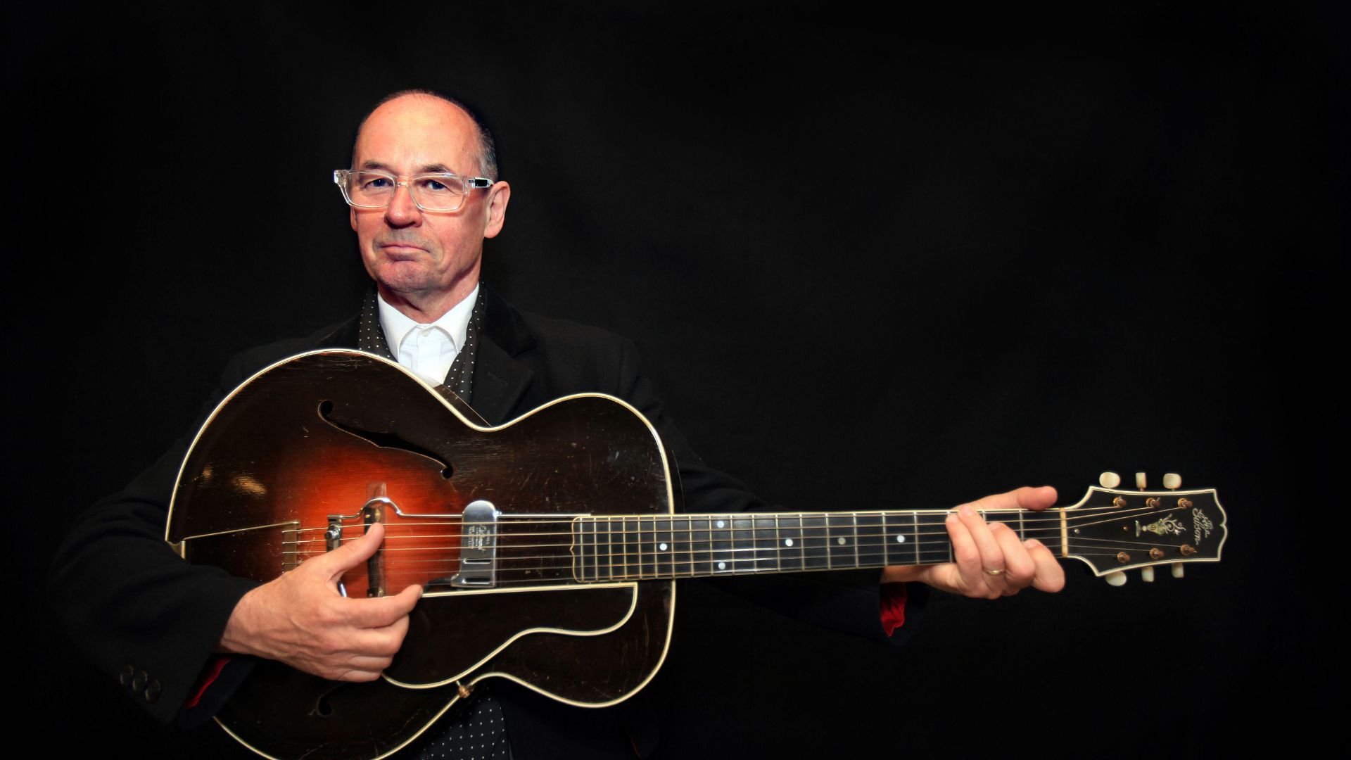 Andy wearing a black suit and holding a guitar, black background