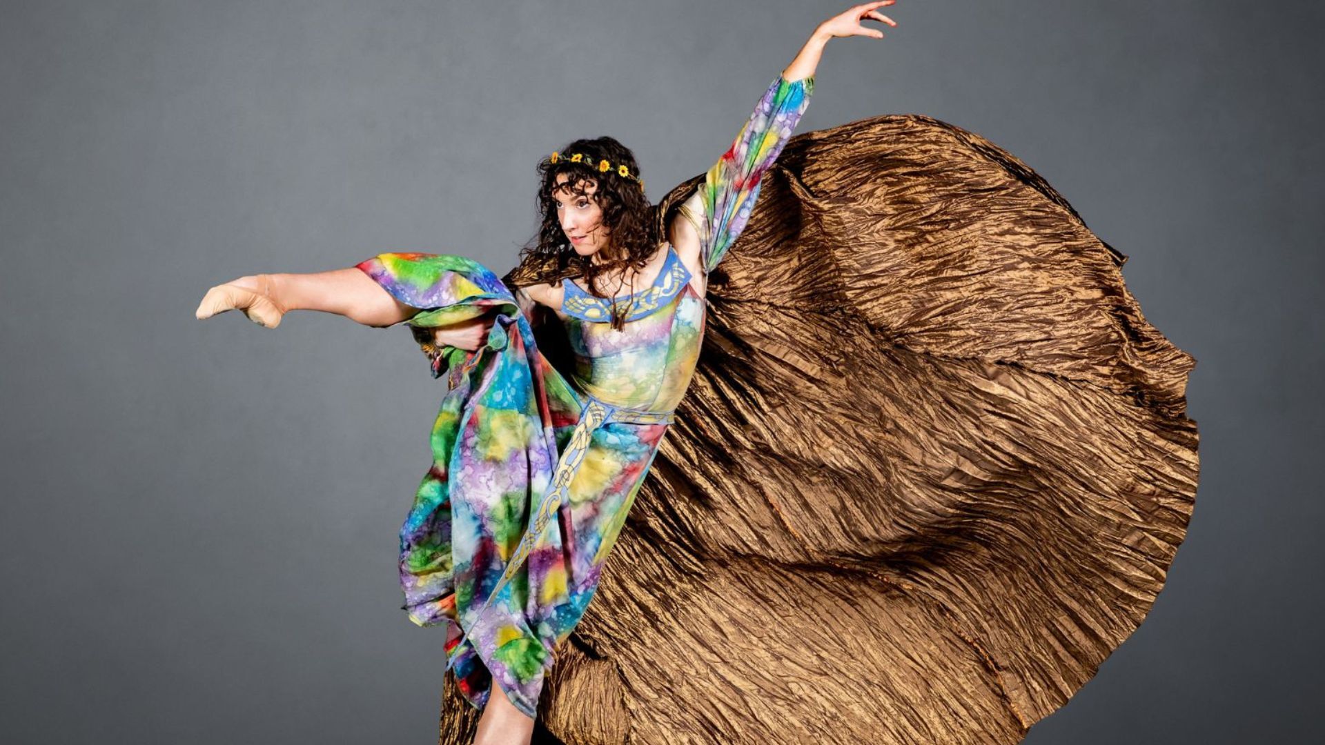 Woman in colourful dress and large brown cloak doing a ballet stance