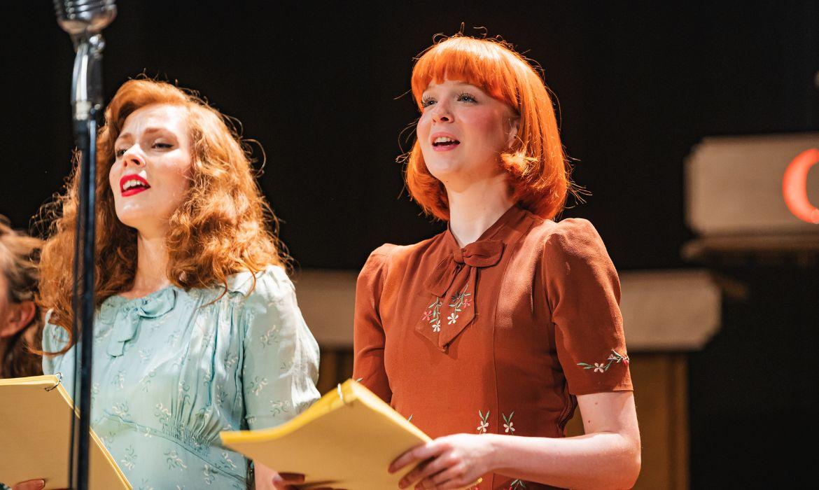 Two women holding scripts and talking or singing into radio studio microphones