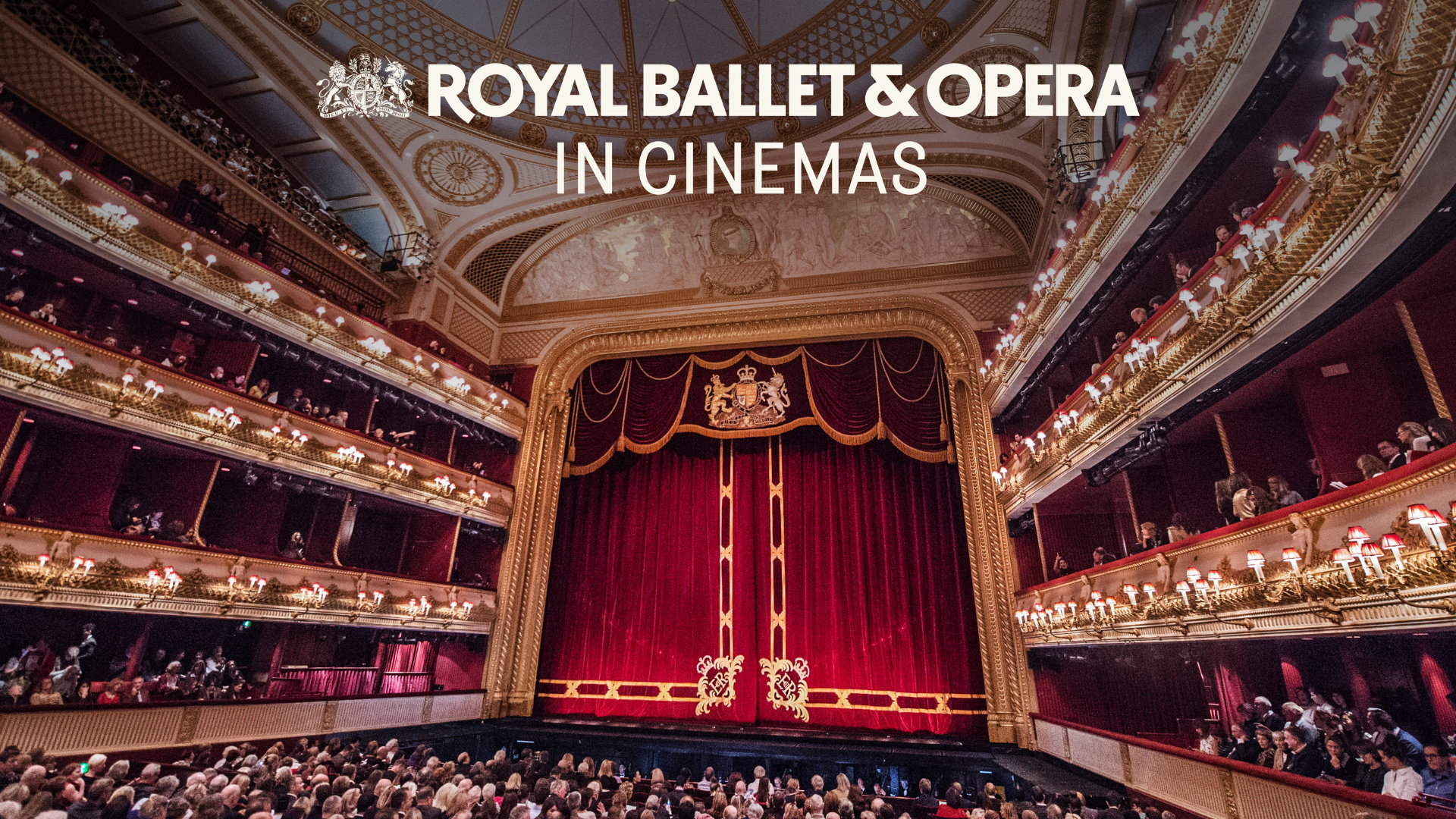 An image of a large and grand theatre with the red curtains drawn and a full audience