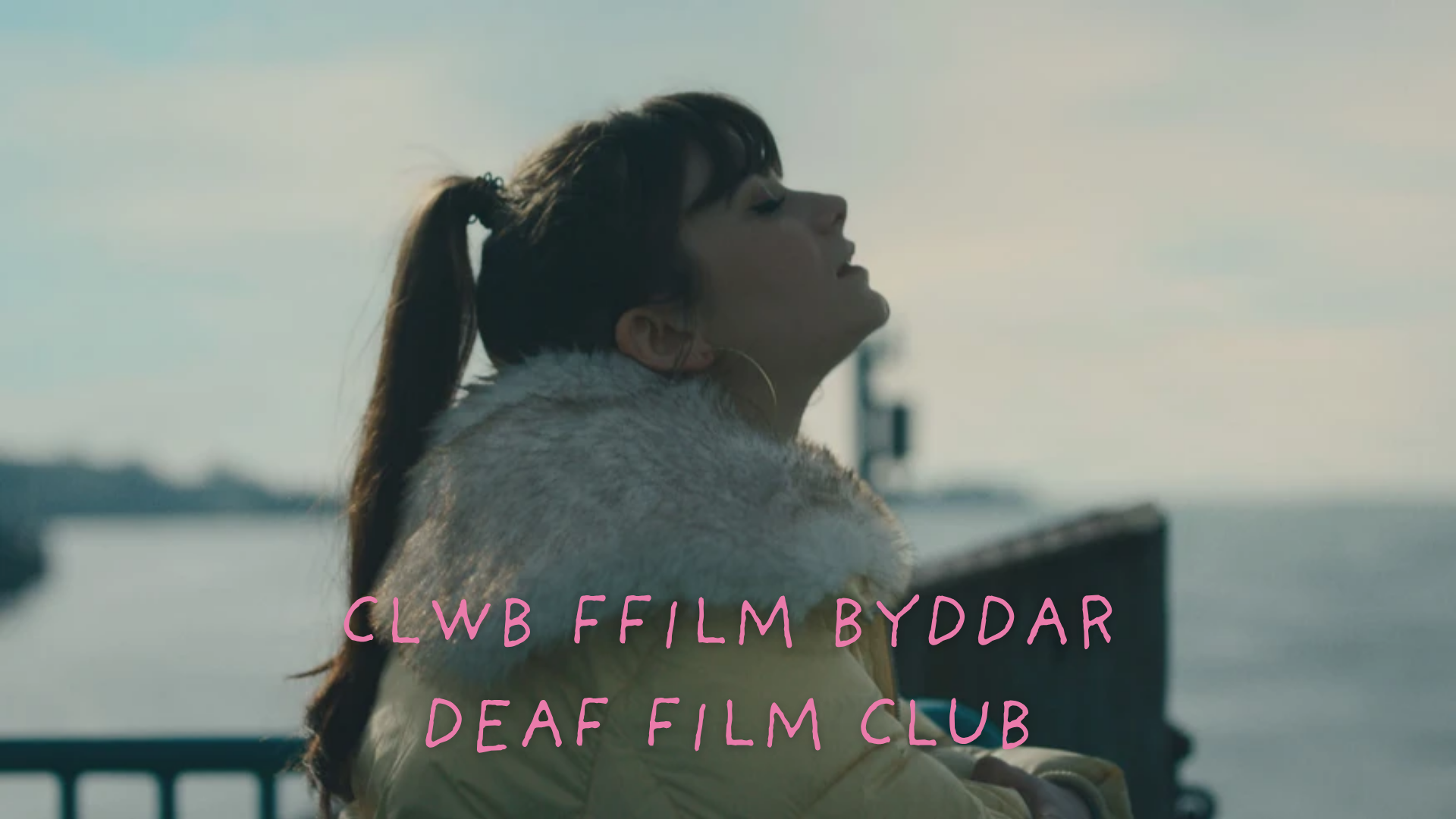 Close up of a young girl sitting by the sea looking up towards the sky with her eyes closed. Deaf Film Club text in a light pink is written along the bottom