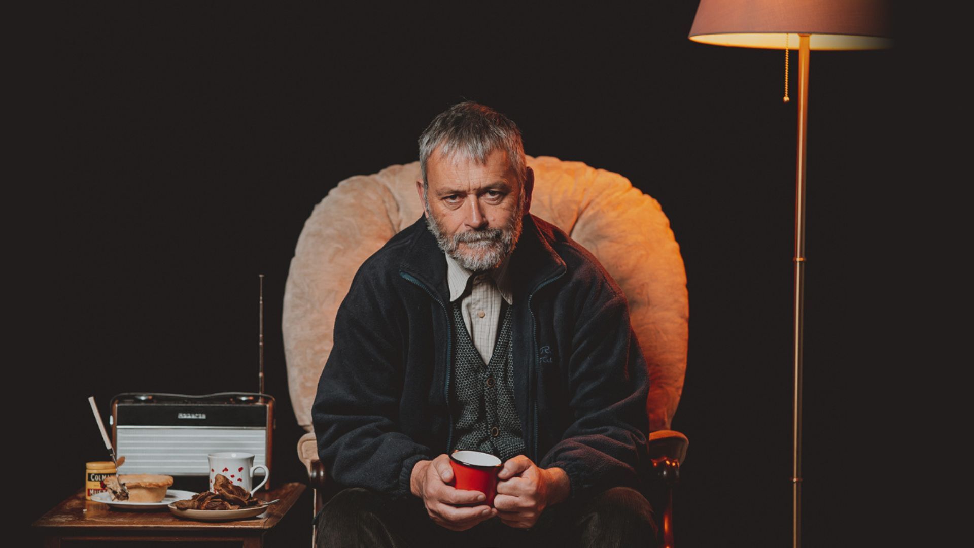 Huw Fyw (played by Tudur Owen) siting in a chair holding mug