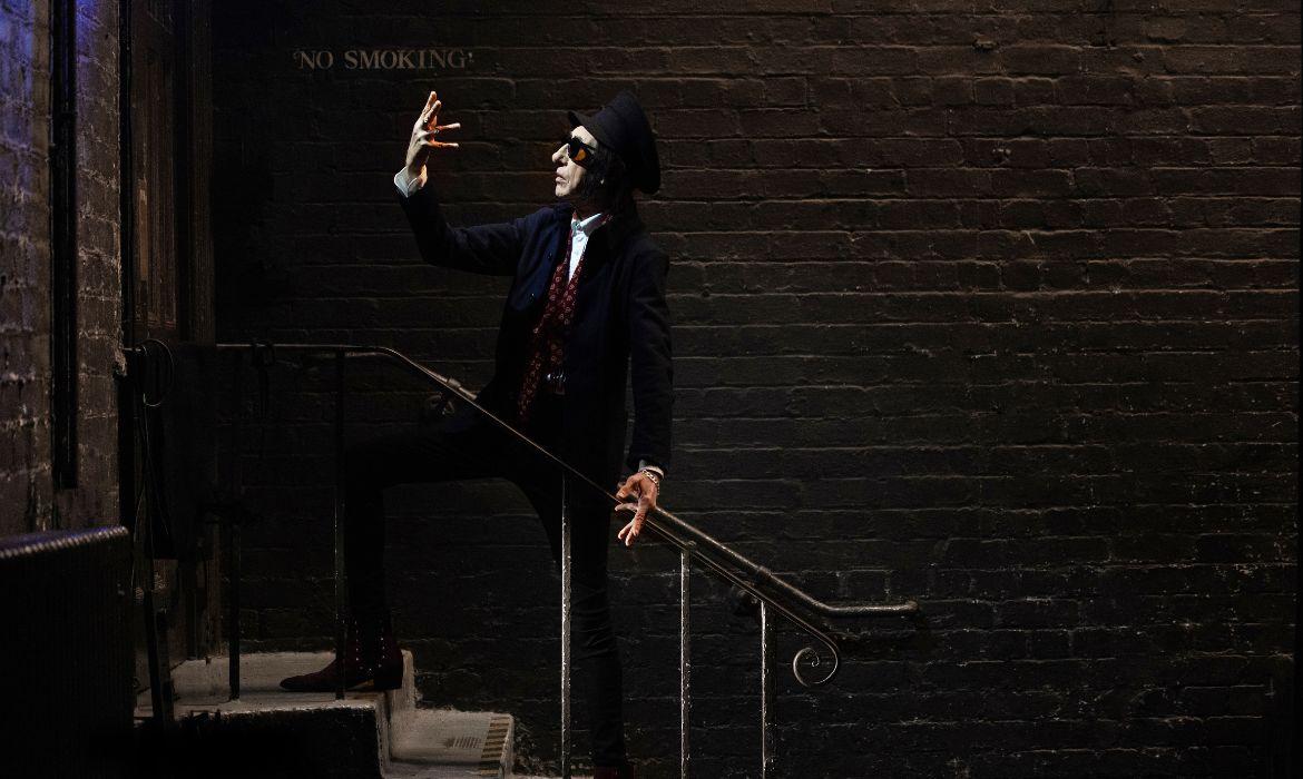 John Cooper Clarke standing on stairs outside a red brick building wearing a dark suit and sunglasses