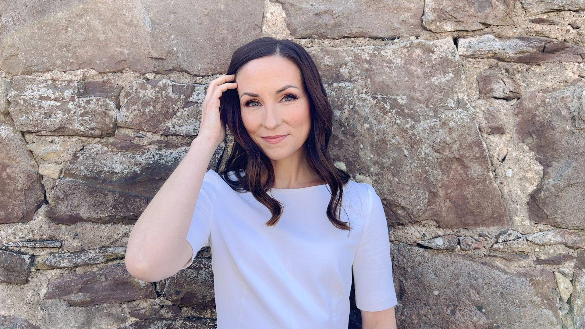 Image of Julie Fowlis wearing a white t-shirt and standing in front of a stone wall