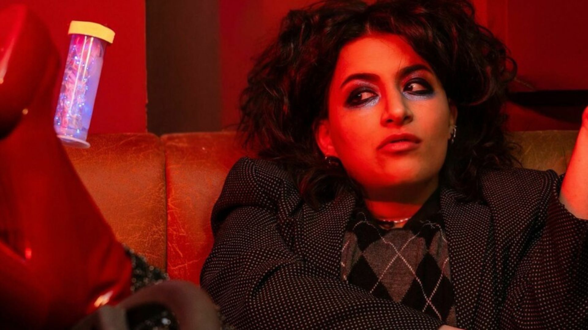 Photo of Leila Navabi slouching on a leather sofa with her feet up, dressed up. Could be a nigh club due to the lighting