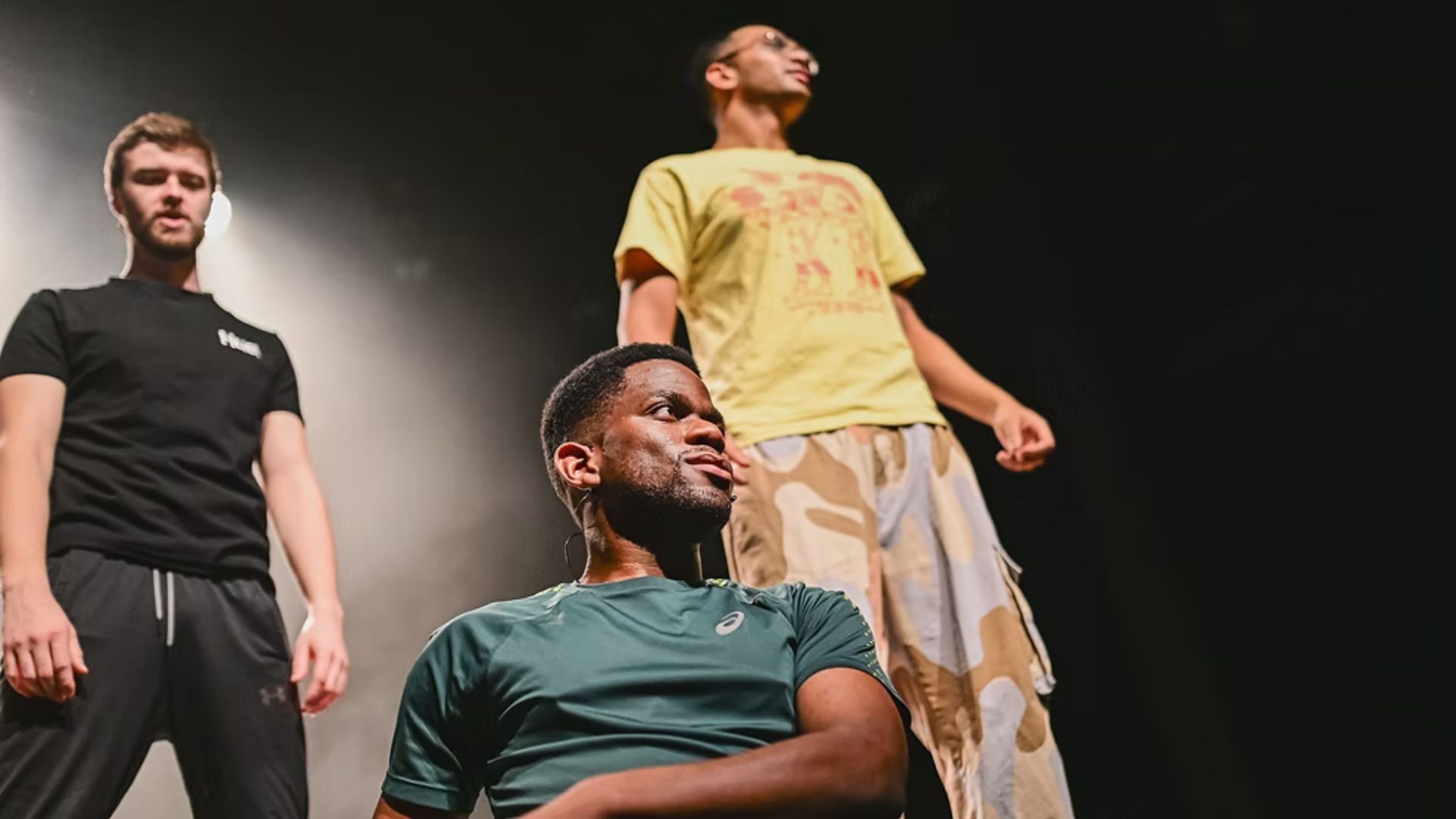 Three men - two standing and one on the floor with dark background and stage lighting