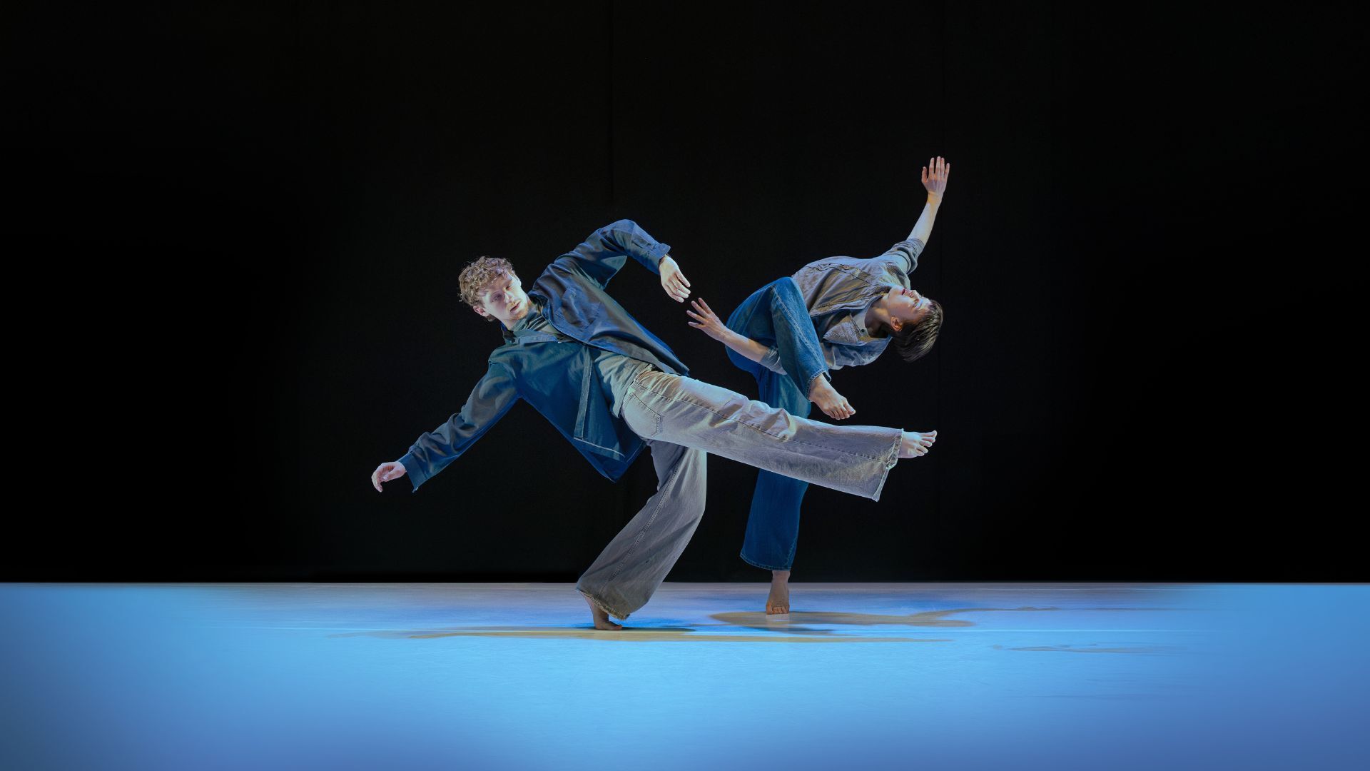 Two people wearing jeans and shirts dancing on stage - black background and blue lit stage