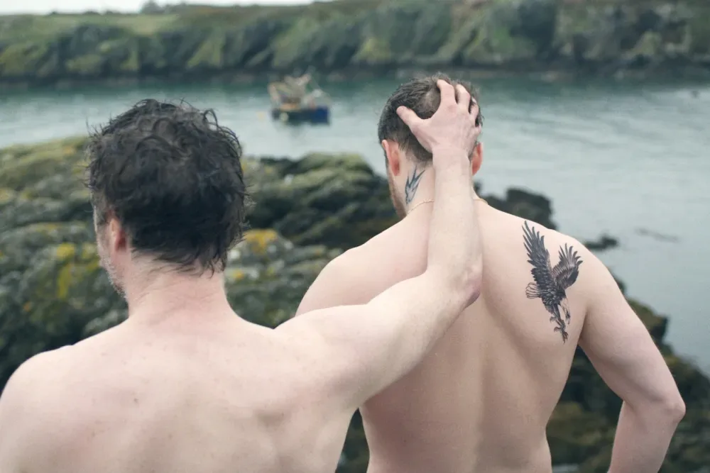 Two topless men from behind walking on rocks on the sea shore
