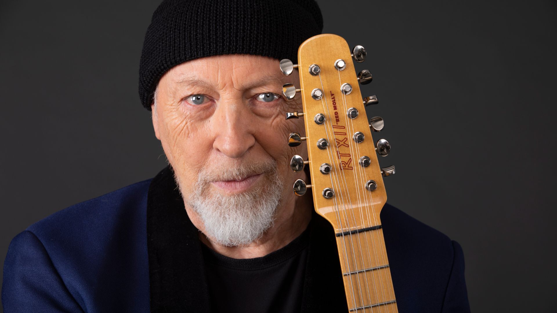 Richard Thompson wearing a blue suit and a black beanie holding his guitar