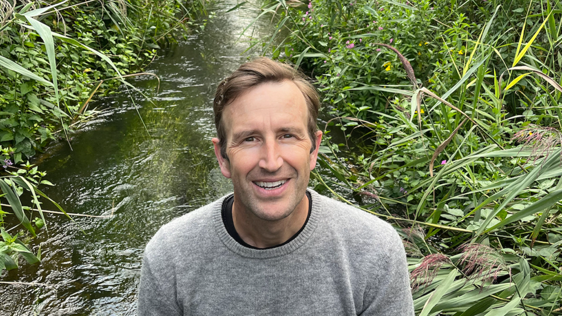 Smiling White man in a grey jumper stood in front of a stream and greenery.
