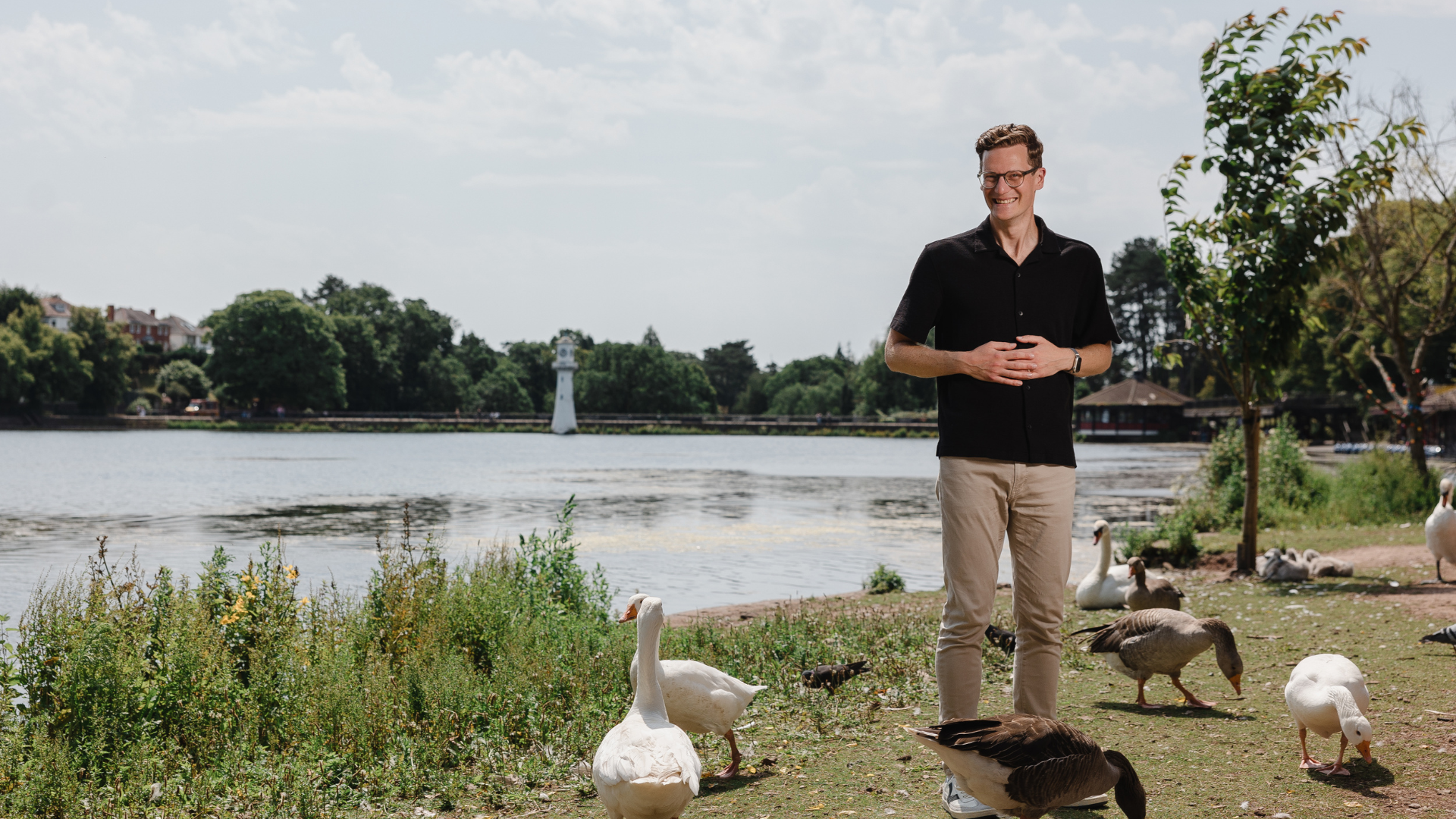 A smiling man in a black shirt and beige pants stands beside a lakeside, surrounded by geese and greenery on a sunny day. A lighthouse and trees are visible across the water.