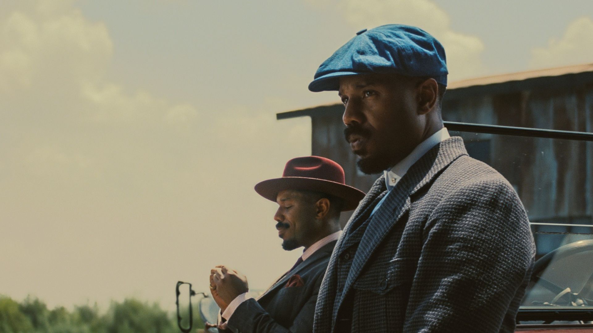 Two men in suits and hats standing near a car with a field and shed in the backgorund