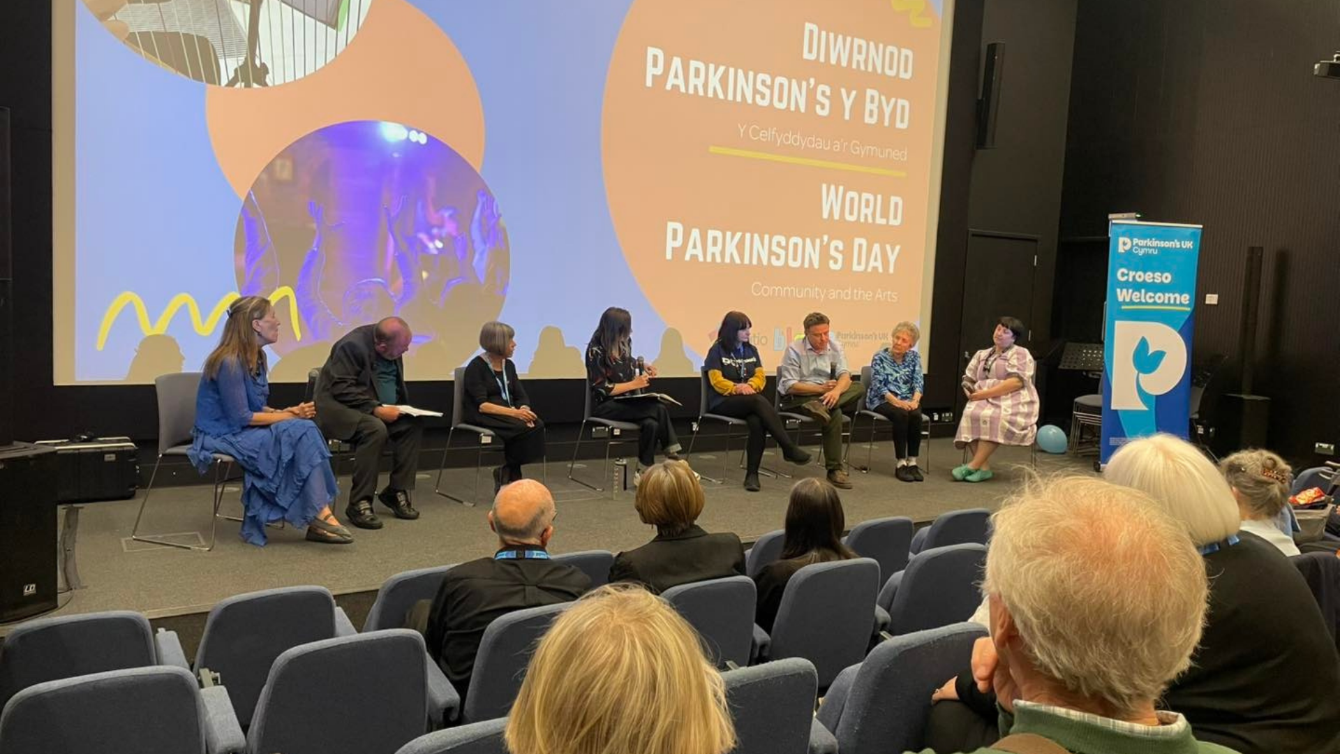 Panel of 8 people sat in discussion on stage in front of a big screen for World Parkinsons Day