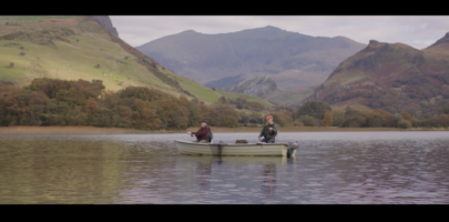 Two men sat in a small fishing boat on a lake surrounded by mountains