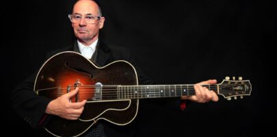 Andy wearing a black suit and holding a guitar, black background