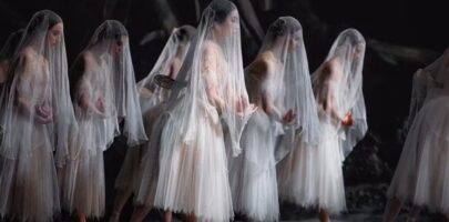 A group of ballerinas in white, ghostly costumes and veils perform a synchronized scene on stage.