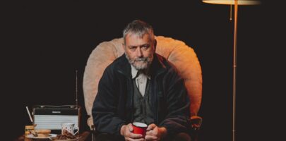 Huw Fyw (played by Tudur Owen) siting in a chair holding mug