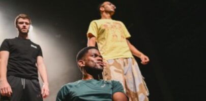 Three men - two standing and one on the floor with dark background and stage lighting