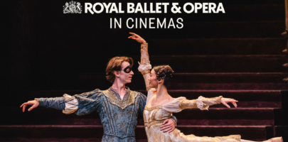 Two ballet dancers a male in blue wearing a mask and a woman in light clothing facing each other, he has his arm around her waist