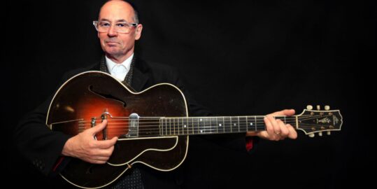 Andy wearing a black suit and holding a guitar, black background