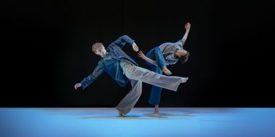 Two people wearing jeans and shirts dancing on stage - black background and blue lit stage