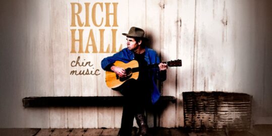 Rich Hall sitting on a bench holding his guitar