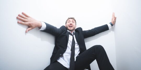 Seann Walsh wearing a suit an tie leaning back against a white wall as if he's in shock