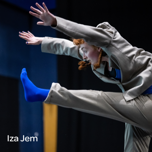 Image of woman wearing a grey suit and bright blue socks dancing with her arms and right leg stretched out in front